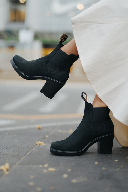 A person wearing a cream-colored skirt and stylish black heeled ankle boots stands outdoors on a sidewalk, with one foot slightly lifted.