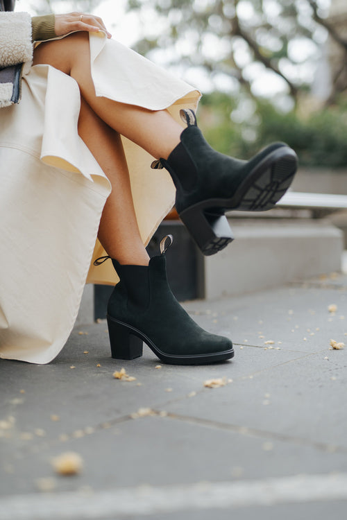 A person sits outdoors with legs crossed, wearing a long cream skirt and black suede ankle boots with chunky heels. Yellow leaves are scattered on the gray pavement around them.