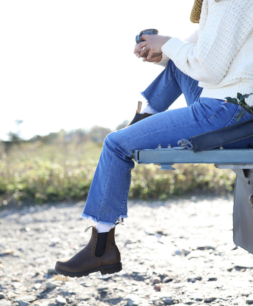 A person wearing blue jeans, brown boots, and a cream sweater sits on the back of a vehicle outdoors, holding a cup, with greenery and rocks in the background.