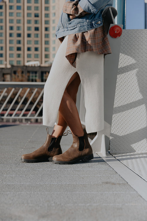 A person in a white knit skirt with a front slit, brown ankle boots, and a layered denim and plaid shirt stands next to a railing outdoors in an urban area.