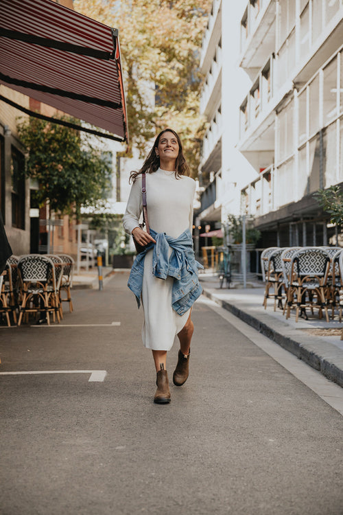 A woman walks confidently down a city street lined with outdoor café tables, wearing a white dress, brown boots, and a denim jacket tied around her waist. Sunlight filters through trees and buildings in the background.