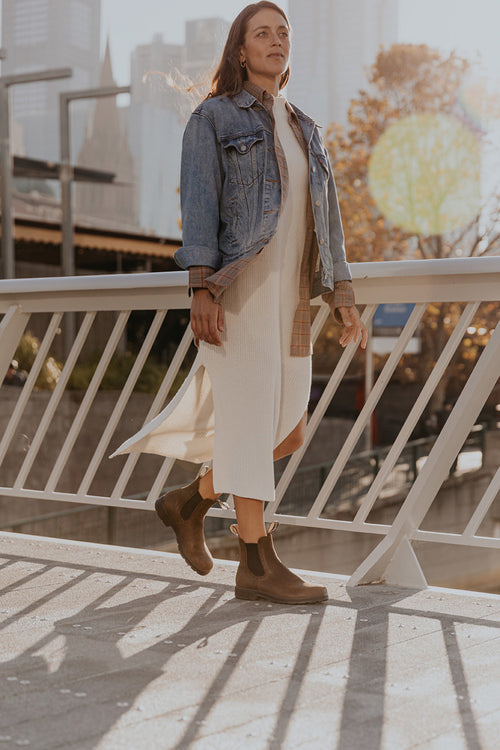 A woman walks outdoors on a sunny day, wearing a white knit dress, denim jacket, brown boots, and a neutral shirt. Autumn trees and city buildings are visible in the background, with sunlight shining through.