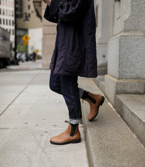 A person wearing cuffed dark jeans, a navy coat, and tan Chelsea boots steps up onto a city sidewalk curb. The upper part of their body and face are not visible.
