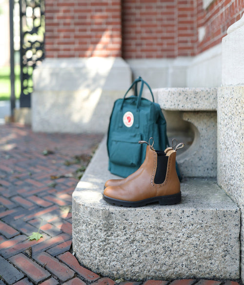 A pair of tan Chelsea boots sits on a stone step next to a green backpack, with a brick building and walkway in the background.