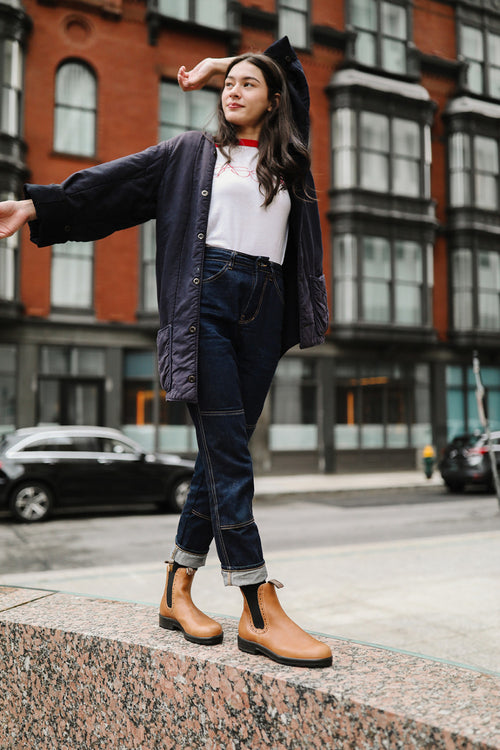 A woman in a black beret, navy coat, white shirt, and rolled-up jeans stands on a stone ledge with one arm raised, smiling. She wears tan boots and is outdoors with buildings and parked cars in the background.