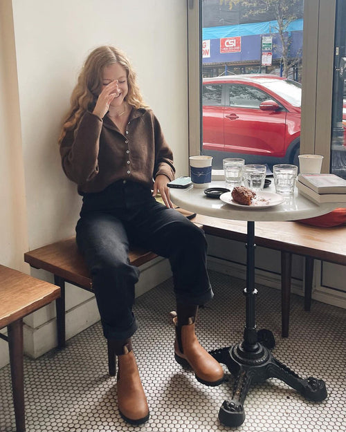 A woman with long wavy hair sits at a small café table by a window, smiling with her hand near her face. On the table are cups, glasses, a slice of cake, and a book. A red car is parked outside.