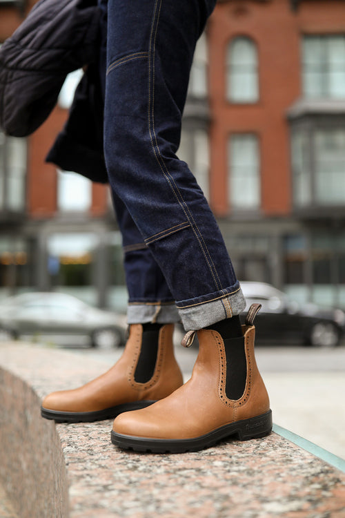 A person wearing cuffed blue jeans and tan Chelsea boots stands on a stone ledge in an urban area, with parked cars and a red brick building blurred in the background.