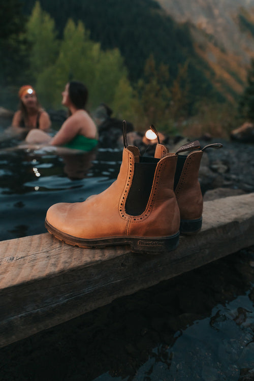 A pair of tan boots sits on a wooden plank near a rocky hot spring, with two women relaxing in the water and wearing headlamps in the blurred background, surrounded by trees and mountains.