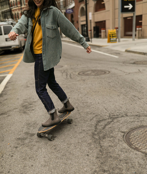 A person in a gray jacket and yellow sweater rides a skateboard on a city street, smiling and balancing with arms outstretched. Cars and buildings are visible in the background.