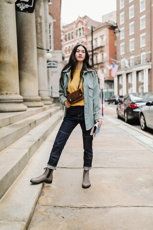 A woman stands confidently on a city sidewalk, wearing a light blue jacket, yellow top, dark jeans, ankle boots, and a brown crossbody bag. She holds a book and looks forward with a neutral expression.