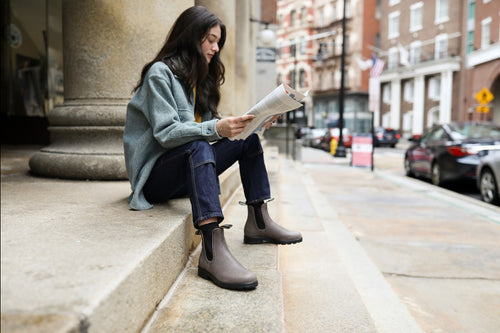 A woman with long dark hair sits on stone steps outside, reading a newspaper. She wears a light green jacket, dark jeans, and brown boots. Buildings and cars are visible in the background.