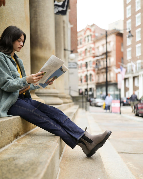 A person wearing a green jacket, blue jeans, and brown boots sits on stone steps outside, reading a newspaper with buildings and street activity in the background.