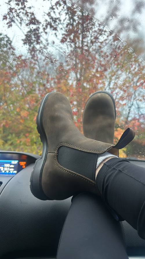 A person wearing brown Chelsea boots and black leggings rests their feet on a car dashboard. Autumn-colored trees and raindrops on the windshield are visible outside.