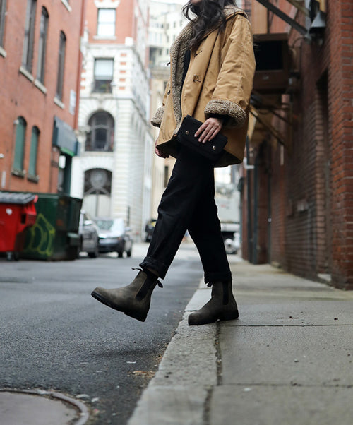 A person wearing a tan jacket, black pants, and brown boots walks along a city sidewalk, holding a black clutch. The background features brick buildings, parked cars, and a red dumpster.