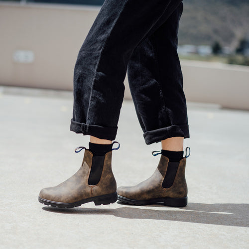 A person wearing cuffed black pants and brown ankle boots with black elastic sides walks outdoors on a sunny day.