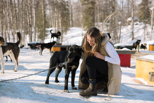 A woman kneels on snowy ground, smiling and petting a black dog on a chain. Several other dogs and colorful doghouses are visible in the background among snow-covered trees.