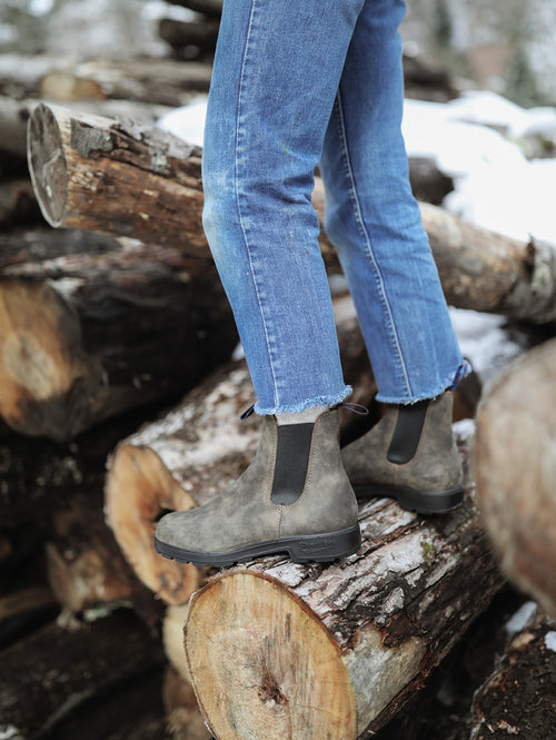 A person wearing blue jeans and brown Chelsea boots stands on a stack of cut logs outdoors, with snow visible in the background.