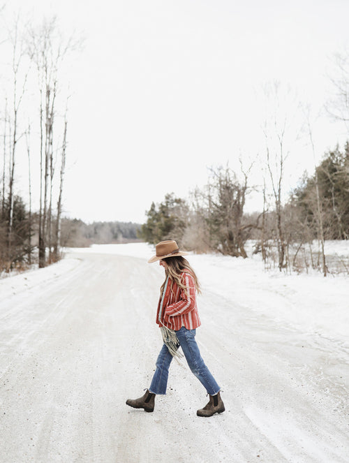 A woman wearing a hat, striped shirt, jeans, and boots walks across a snowy, rural road surrounded by bare trees and winter landscape.