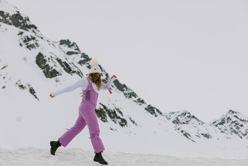 A person in a purple snowsuit and white hat walks on snow with mountains in the background under an overcast sky.