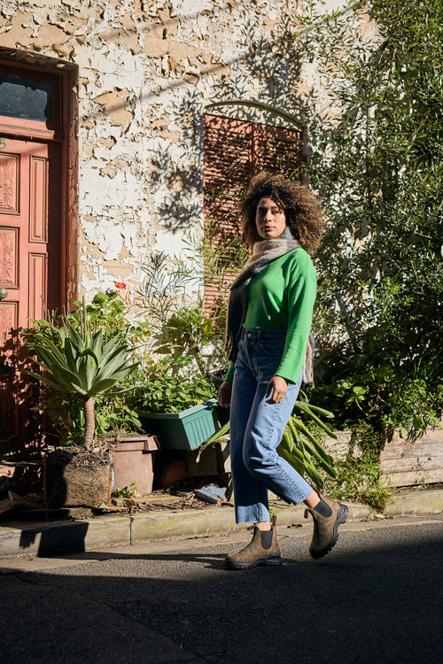 A person with curly hair wearing a green sweater, scarf, jeans, and boots walks past potted plants and a weathered wall with a red door and window in bright sunlight.