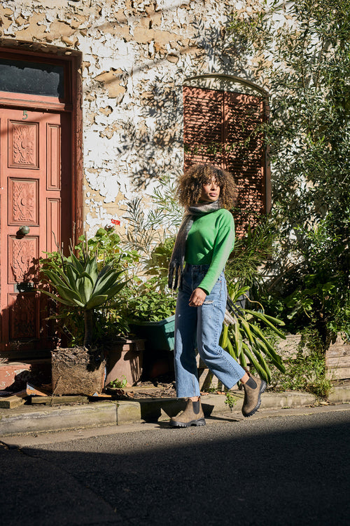 A woman with curly hair wearing a green sweater, blue jeans, and boots walks past a rustic building with a wooden door and potted plants in bright sunlight.