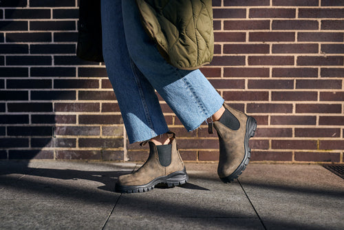 A person wearing blue jeans, olive green quilted coat, and brown Chelsea boots is walking on a sunlit sidewalk in front of a brick wall. Only the legs and feet are visible.