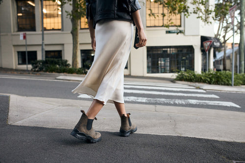 A person wearing a flowing white skirt, a black jacket, and brown ankle boots is walking across a city street at a crosswalk. Trees and buildings are visible in the background.