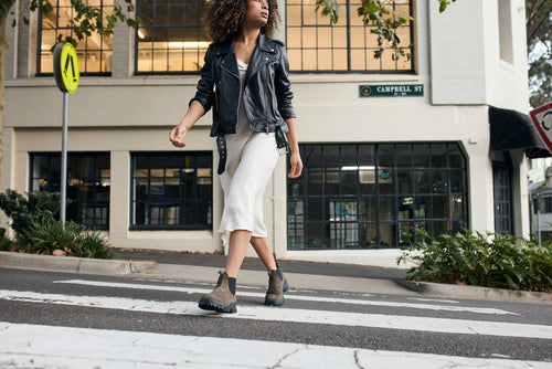 A woman wearing a black leather jacket, white dress, and boots walks confidently across a crosswalk on a city street lined with buildings and trees.