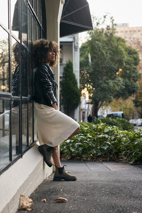 A person with curly hair, wearing a black leather jacket, white skirt, and boots, leans against a building with large windows. Lush greenery and city buildings are visible in the background.