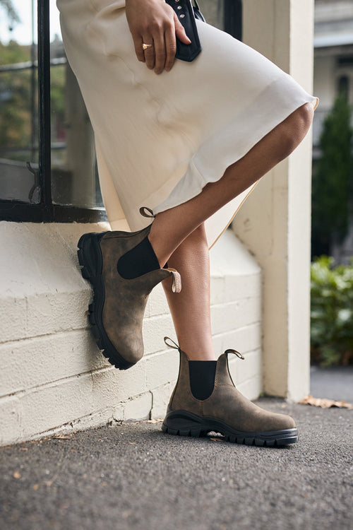 A person in a cream skirt leans against a white wall outdoors, wearing brown Chelsea boots with black elastic sides and chunky soles. The background shows greenery and a building.