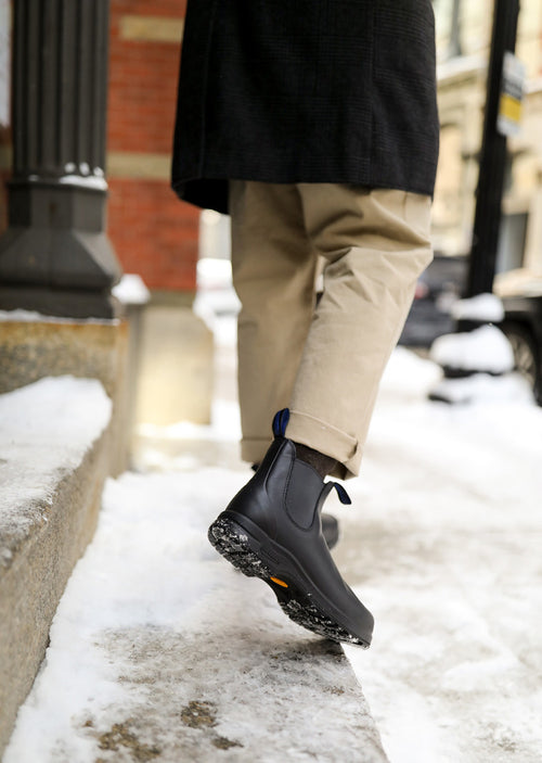 A person wearing tan pants and black boots steps onto a snowy sidewalk curb in an urban setting, with slush and snow on the ground and buildings visible in the background.
