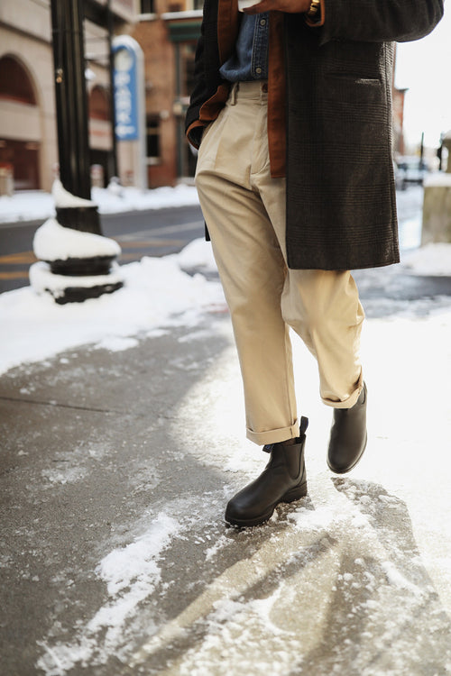 A person wearing light khaki pants, black boots, a blue shirt, and a dark coat walks on a snowy city sidewalk. Snow is on the ground and nearby surfaces, with buildings and a street in the background.