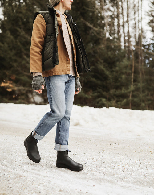 A person walks outdoors on a snowy path, wearing cuffed jeans, black boots, a brown jacket with a black vest, gray knit gloves, and a beanie. Evergreen trees and snow are visible in the background.