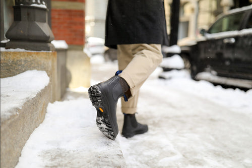 A person wearing beige pants and black boots walks on a snowy city sidewalk, lifting one foot to reveal the boots textured sole. Snow covers the ground and nearby steps.