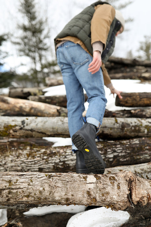 A person in blue jeans, boots, a green vest, and a brown jacket climbs over a stack of snow-dusted logs outdoors, with trees and overcast sky in the background. The photo is taken from behind and below.