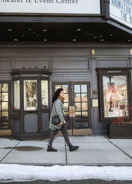 A woman wearing a green quilted coat, black pants, and boots walks past the entrance of a theater and event center on a snowy sidewalk. She carries a black bag and looks ahead as she walks.