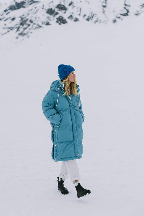 A woman in a blue puffer coat, blue beanie, white pants, and black boots walks on snow with snowy mountains in the background.