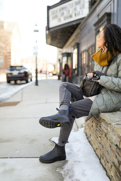 A woman wearing a green coat, mustard scarf, and black boots sits on a stone ledge by a snowy sidewalk, looking toward a theater marquee on a city street.