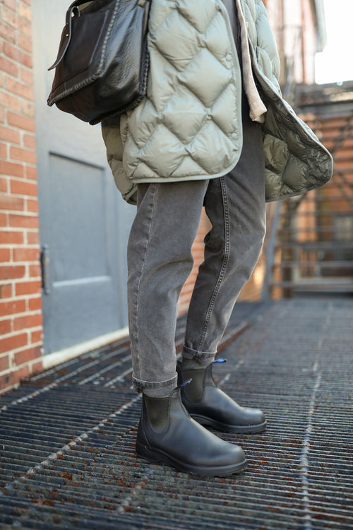 A person stands on a metal grate wearing cuffed gray jeans, black boots, a light quilted coat, and carrying a black leather bag. A brick wall and blue door are in the background.