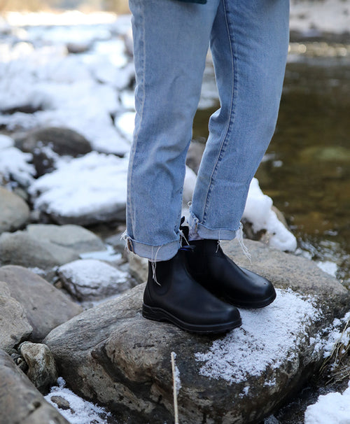 A person wearing light blue jeans and black boots stands on a snow-dusted rock beside a stream, surrounded by rocks with patches of snow.
