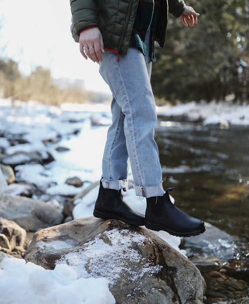A person wearing cuffed jeans, black boots, and a green jacket stands on a snowy rock beside a flowing river in a winter landscape with trees in the background.
