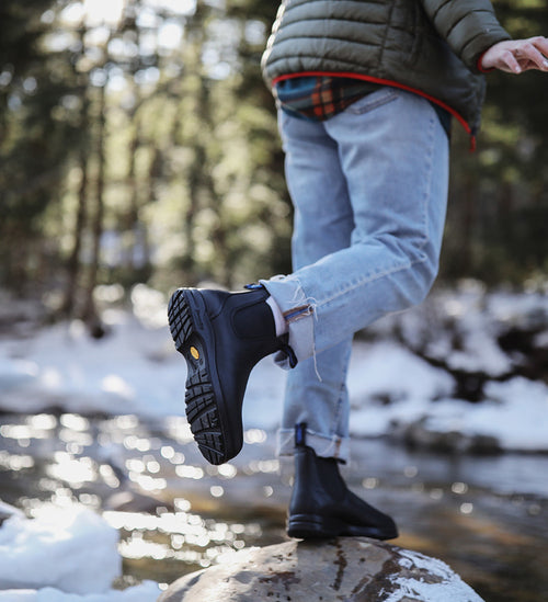 A person wearing ripped jeans, a green jacket, and black boots balances on a rock by a snowy, flowing creek in a forest.