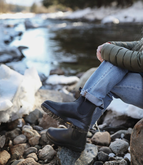 A person in blue jeans, black boots, and a green jacket sits on rocks by a snowy riverbank, with their legs crossed and hands resting on their knee. The background shows snow, rocks, and water.