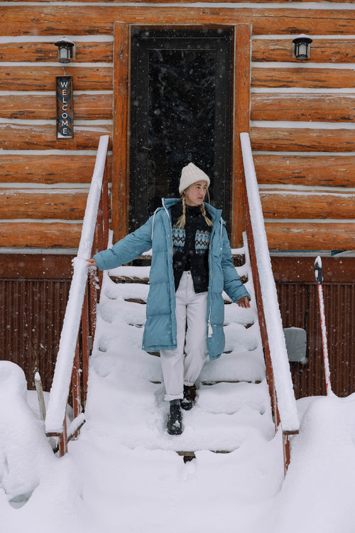 A woman in a blue winter coat, white pants, and a knit beanie stands on snow-covered steps outside a log cabin, with snow falling around her. A vertical WELCOME sign hangs by the door.