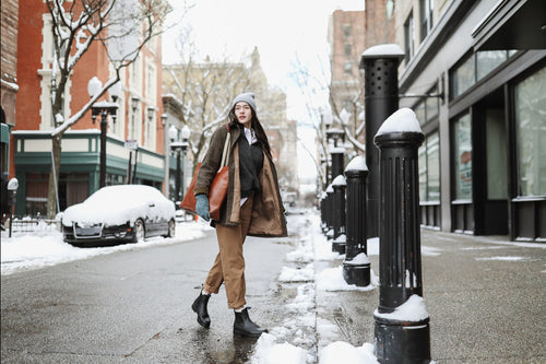 A woman wearing a beanie, scarf, and winter coat walks on a snowy city sidewalk with snow-covered cars, trees, and buildings in the background. She carries a large brown bag and looks to the side.