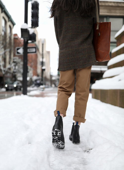 A person walks away on a snowy city sidewalk, leaving footprints. They wear tan pants, a dark coat, black boots, and carry a brown tote bag. Snow is piled on the side, and buildings line the street.