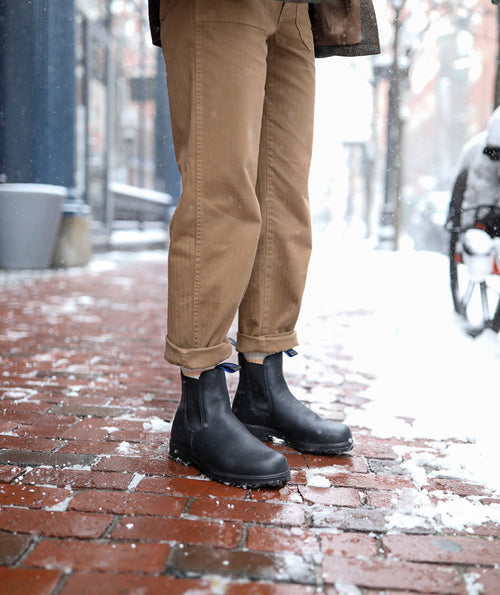 Person wearing tan pants and black boots standing on a snowy brick sidewalk in a city. Snow is falling and lightly covering the ground and nearby parked car. The photo focuses on the lower half of the person’s legs.