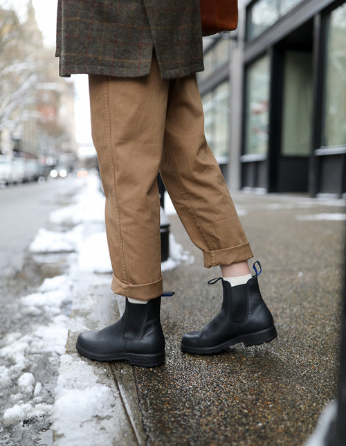 Person standing on a wet, snowy sidewalk wearing cuffed tan pants and black ankle boots; the background shows an urban street with buildings and patches of melting snow.