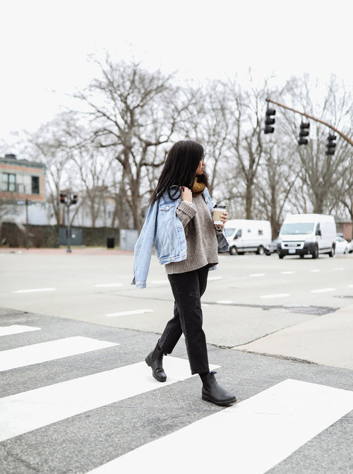 A woman with long dark hair crosses a street at a crosswalk, wearing a gray sweater, black pants, black boots, and a blue denim jacket draped over her shoulders, holding a coffee cup. Trees and vehicles are in the background.