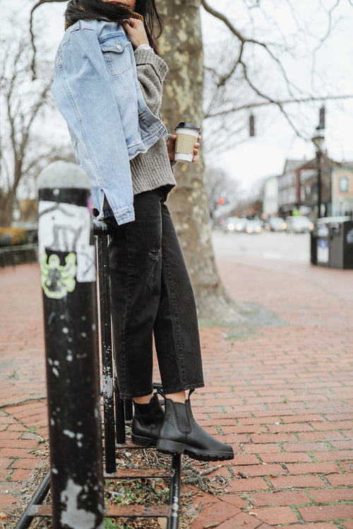 A person in black jeans, black boots, a gray sweater, and a light denim jacket draped over their shoulders holds a coffee cup while leaning against a bike rack on a brick sidewalk with trees and buildings in the background.